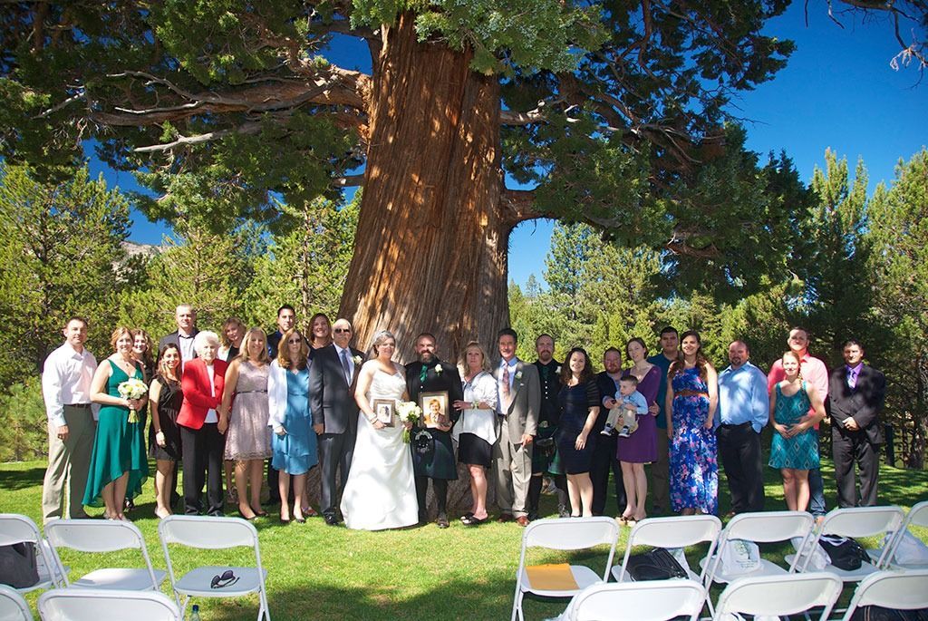 Wedding group photo under a large tree; couple in the center, guests in colorful attire, chairs in front.
