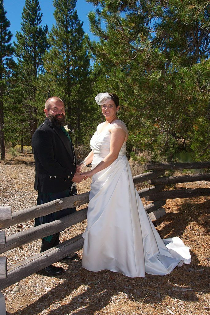 Wedding couple poses by wooden fence in a forest setting. Bride in white dress, groom in kilt.