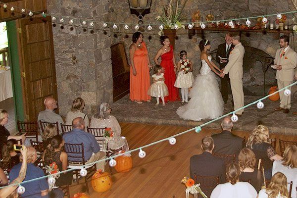 Wedding ceremony indoors; couple holding hands near fireplace, guests seated, string lights overhead.