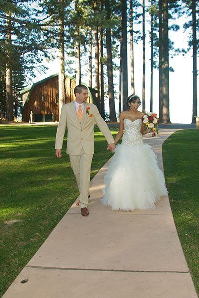Bride and groom walk hand-in-hand on path. Building in background, trees and grass.