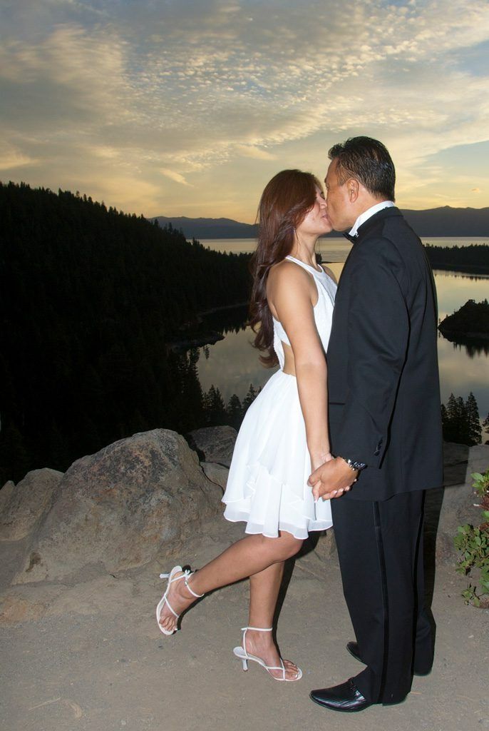 Couple kissing, holding hands on a cliff, overlooking a lake at sunset. The woman is wearing a white dress.