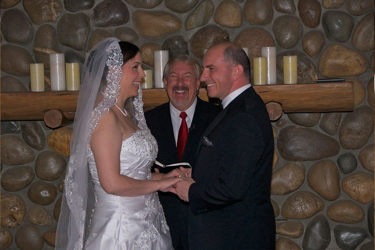 Bride and groom holding hands during wedding ceremony; officiant smiles. Stone wall backdrop, candles.