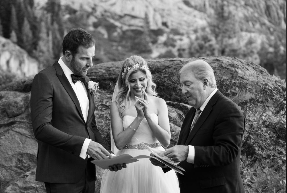 Wedding ceremony: Groom and bride with officiant, exchanging vows outdoors near rocks.