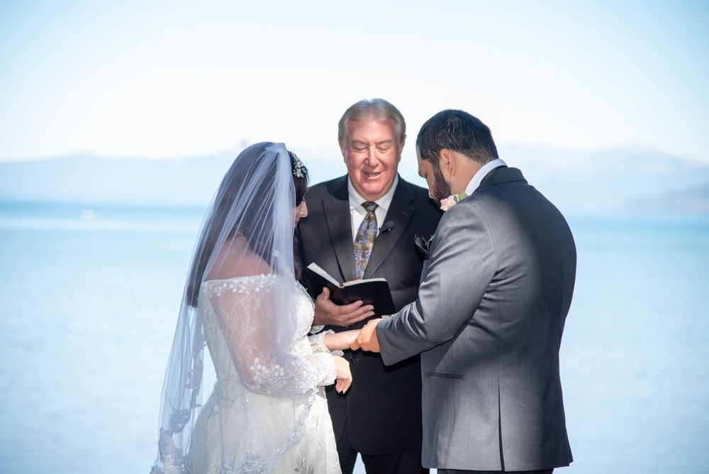 Couple exchanging rings during a wedding ceremony outdoors, officiant present; water in background.