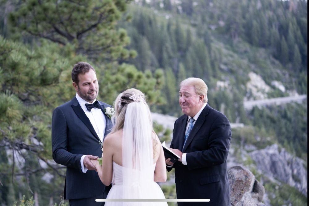 Wedding ceremony outdoors, couple with officiant; mountains in background. Groom in tuxedo, bride in white dress.