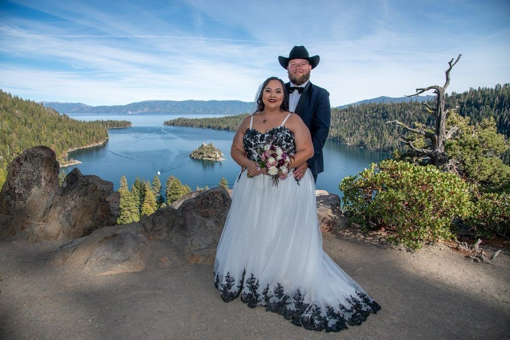 Couple in wedding attire poses by lake, bride in white and black gown, groom in suit with cowboy hat.