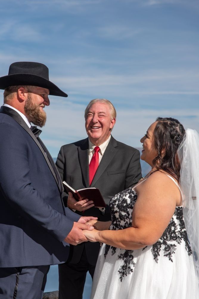 Bride and groom holding hands during outdoor wedding ceremony; officiant laughs, blue sky backdrop.