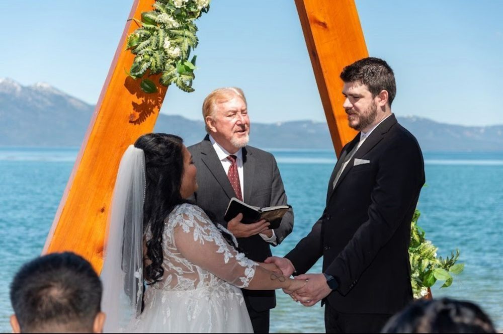 Couple holding hands during wedding ceremony by the water, wooden arch in background.