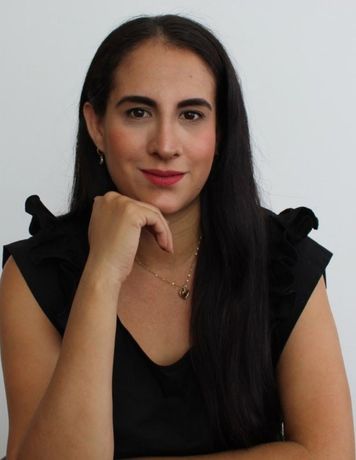Mujer con blusa negra posando con la mano bajo la barbilla sobre un fondo gris claro.