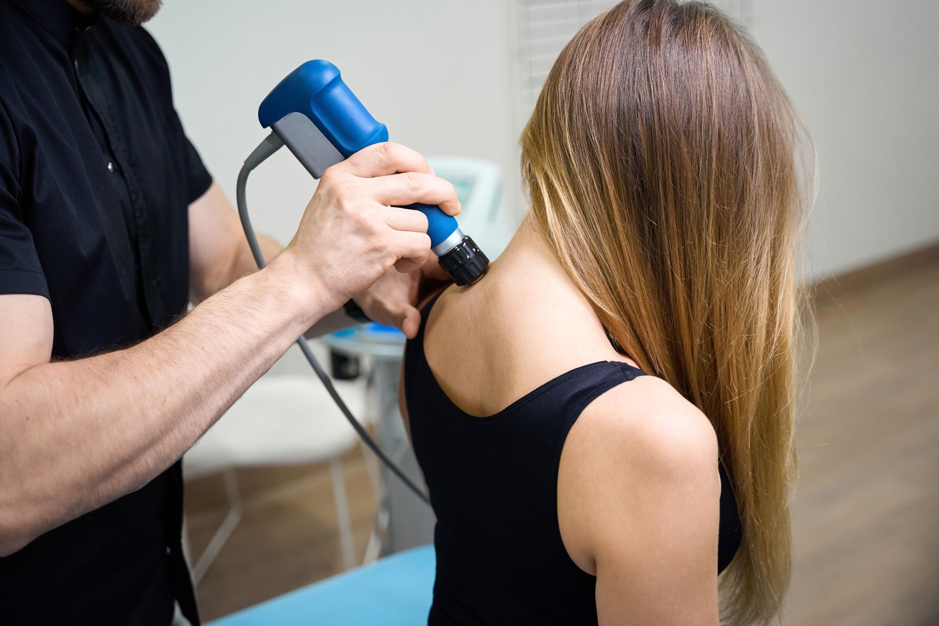 Therapist using a blue device on a woman's shoulder and neck; indoors.