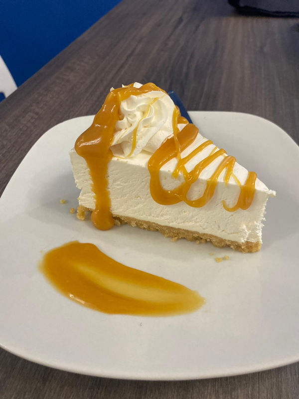 Una fetta di cremosa cheesecake su un piatto bianco, guarnita con panna montata e irrorata con una salsa al caramello.