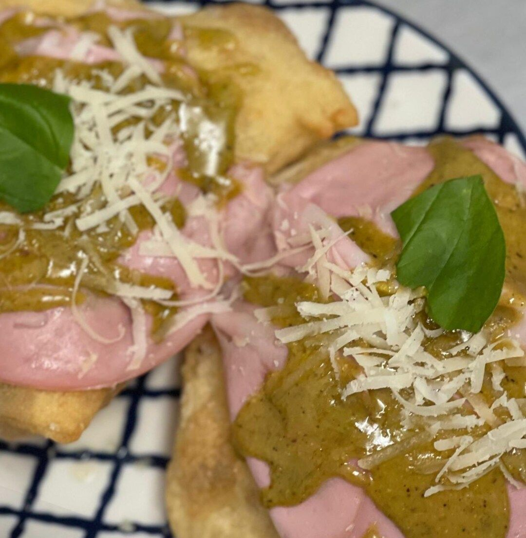 Due pezzi di pasta fritta guarniti con fette di prosciutto, pesto verde, formaggio grattugiato e foglie di basilico fresco.