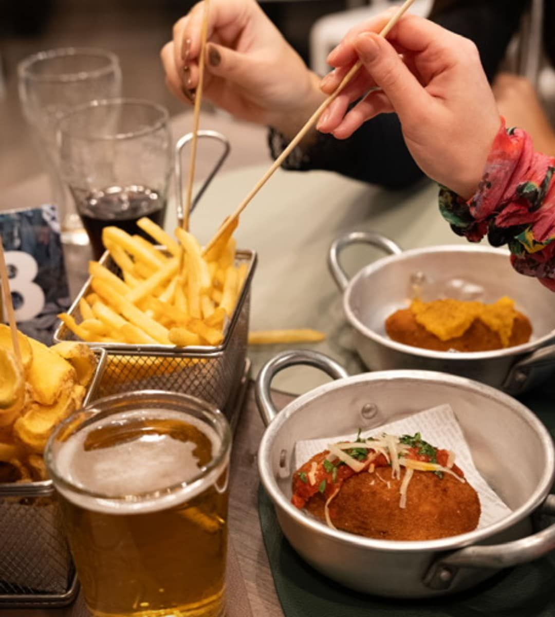 Una persona mangia patatine fritte da un cestino con degli spiedini, accanto a piccole padelle di cibo fritto e di birra.