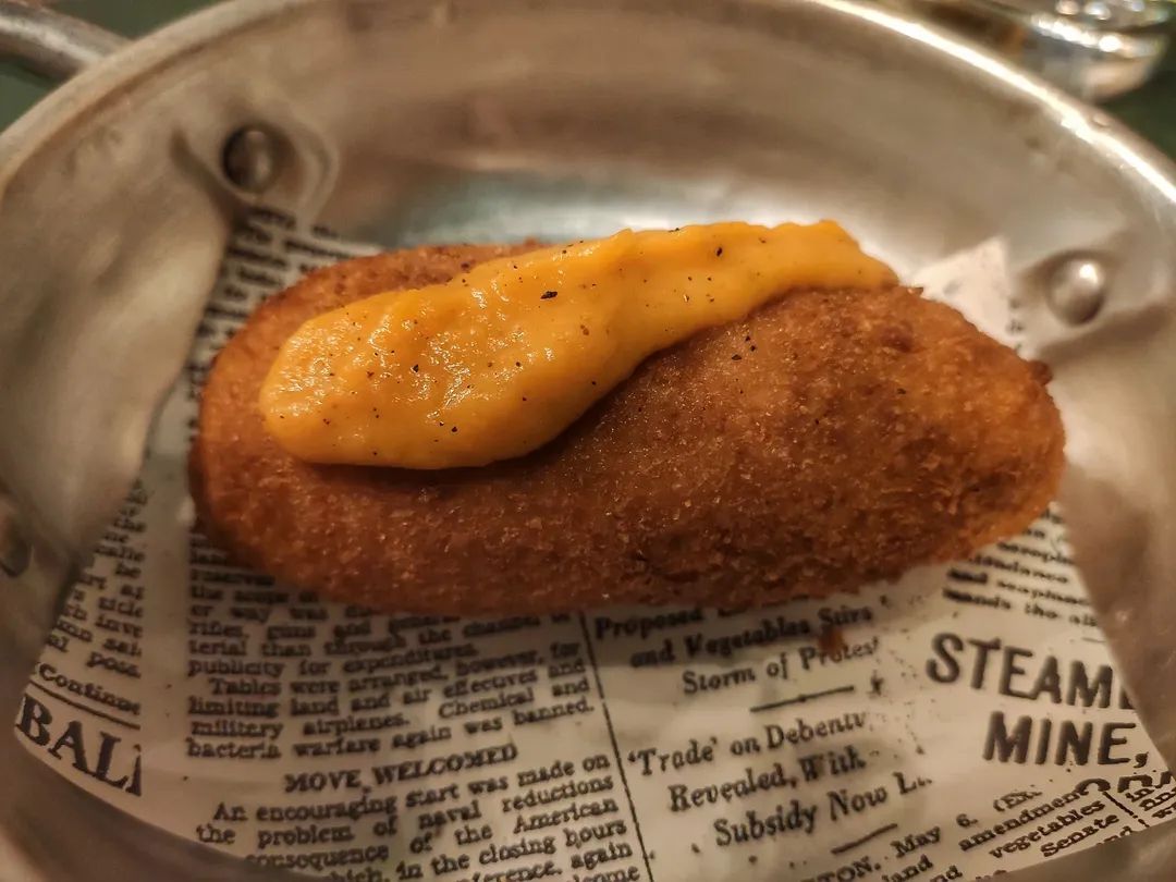 Una crocchetta dorata, guarnita con una cucchiaiata di salsa, servita in una padella di metallo su carta di giornale.