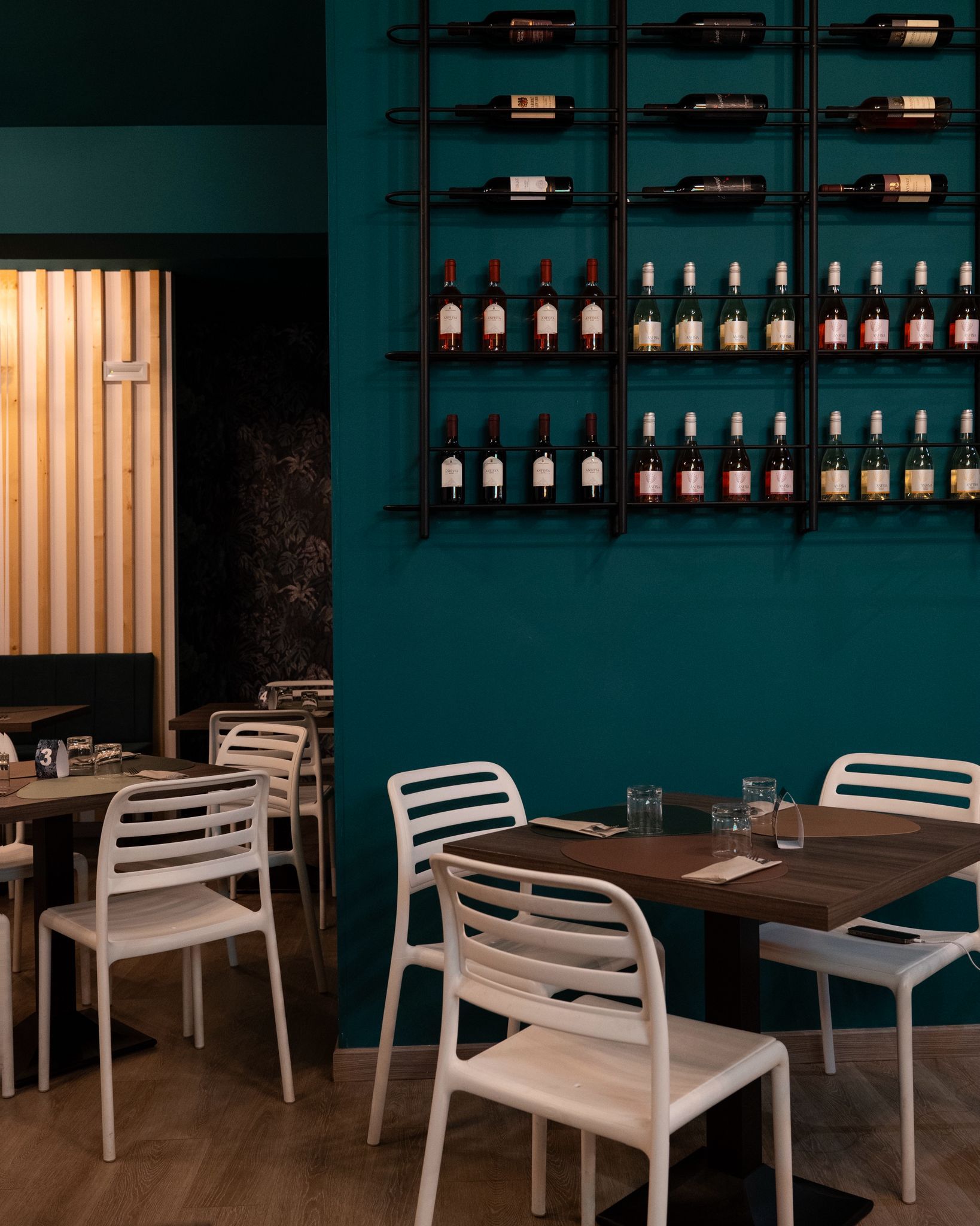 Zona pranzo all'aperto con tavoli e sedie bianche illuminate da luci decorative, situata di fronte a un edificio a più piani.