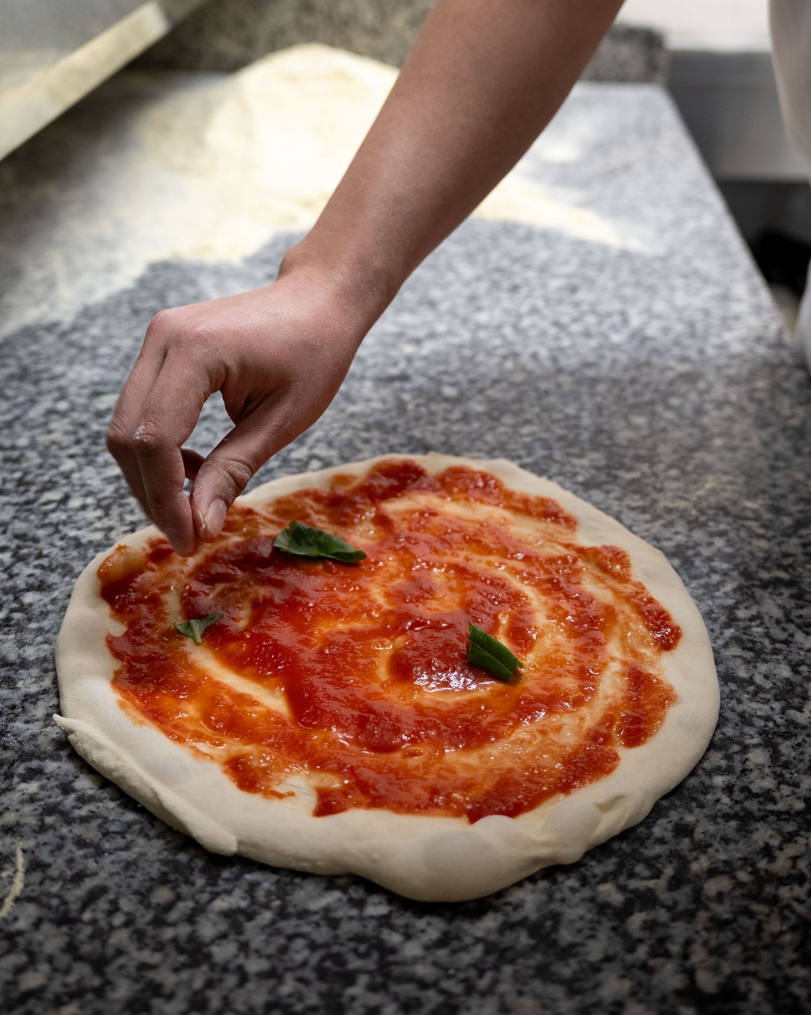Una mano posiziona foglie di basilico su una base di pizza cruda condita con salsa di pomodoro e strisce di mozzarella.