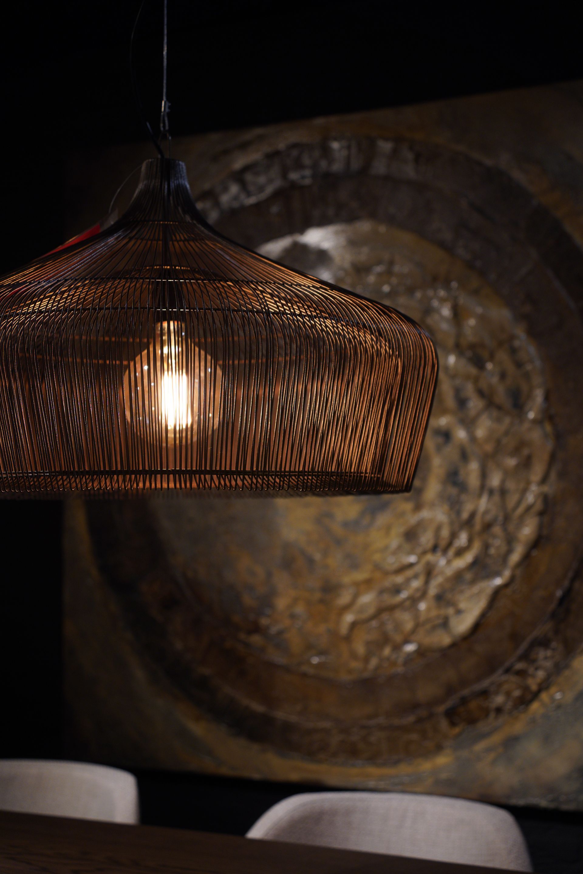 Openwork bronze pendant lamp with exposed bulb, over a table, and a dark abstract painting.