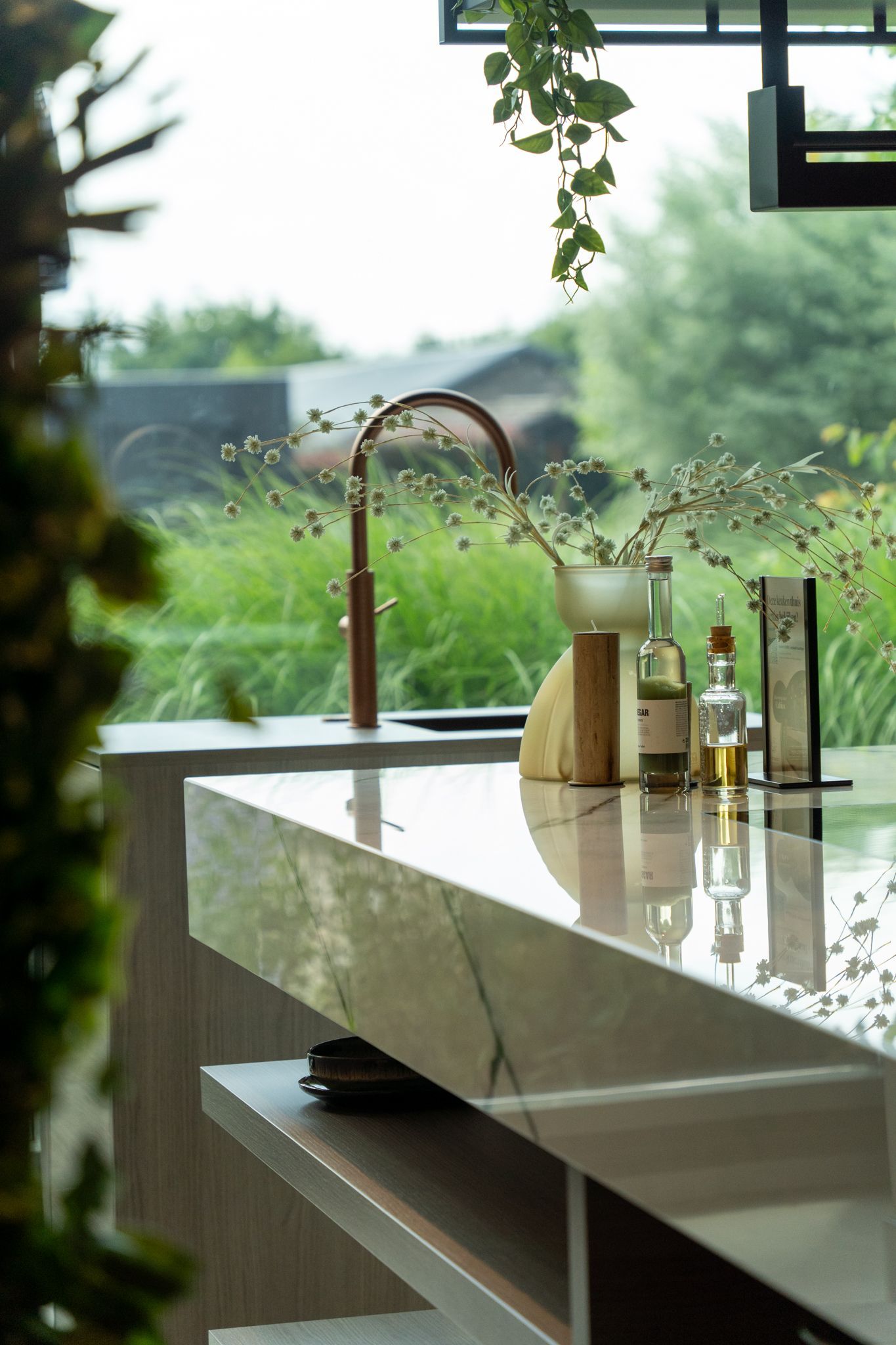 Kitchen counter with marble top, copper faucet, glass bottles, and greenery with a blurred outdoor view.