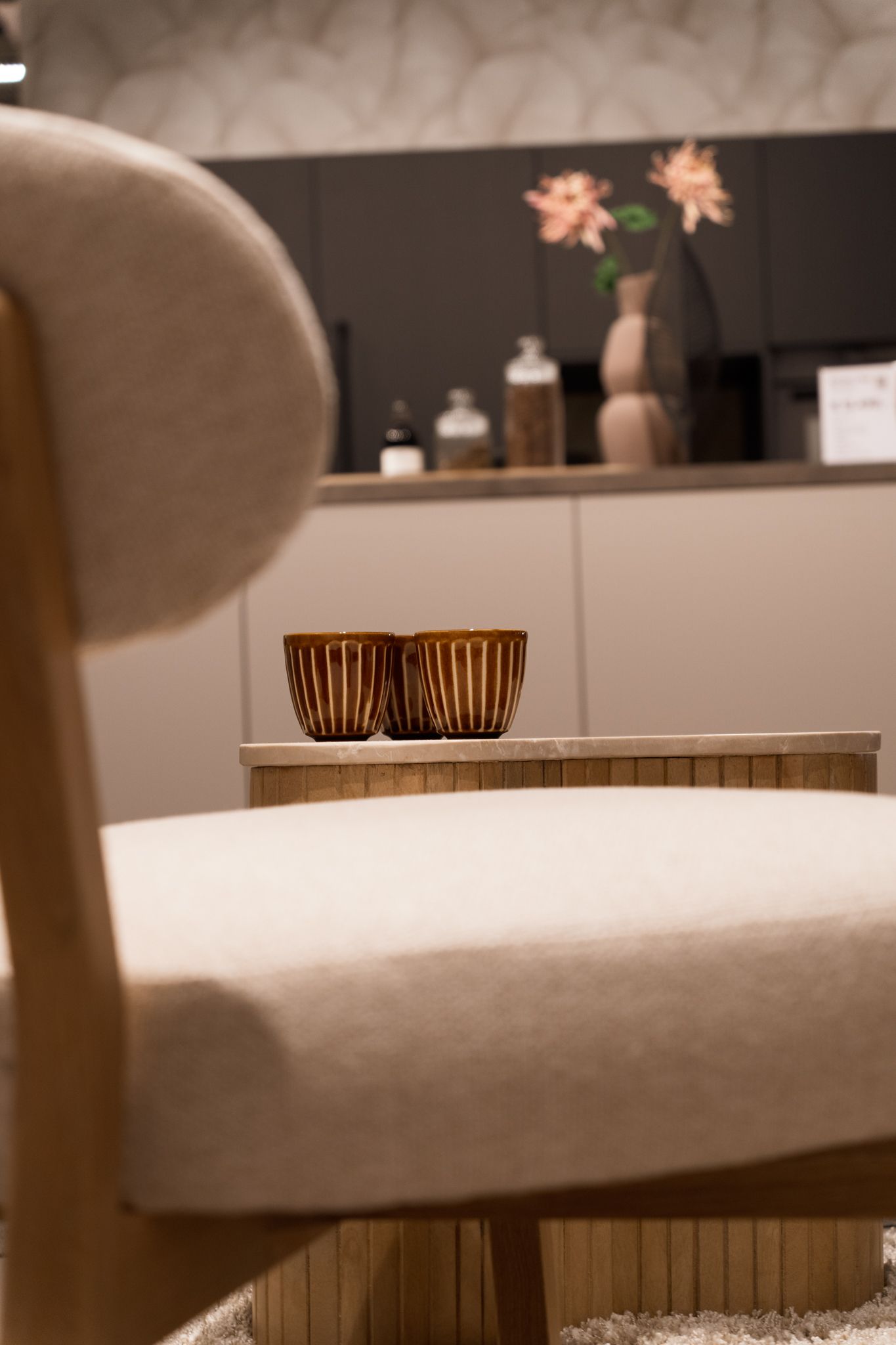 Chair with a small table, holding two decorative amber-colored containers. Background includes a cabinet, flowers, and jars.