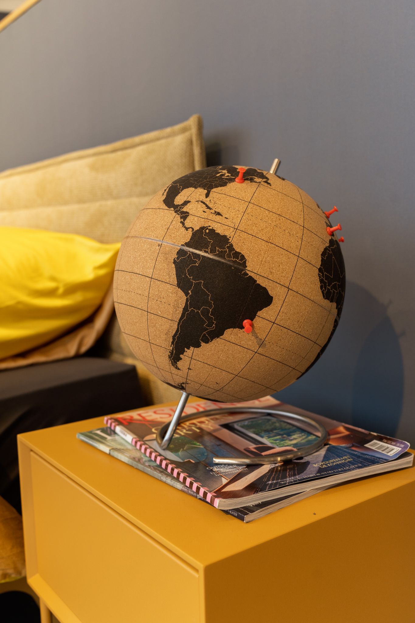 Cork globe with red pins on a yellow bedside table, with magazines.
