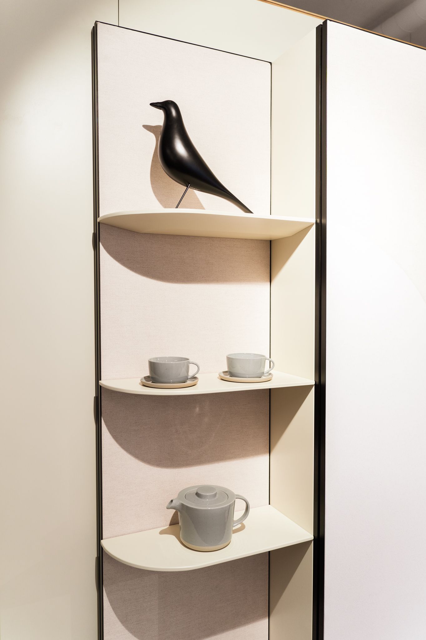 Black bird sculpture and gray teacups on shelves against a white partition.