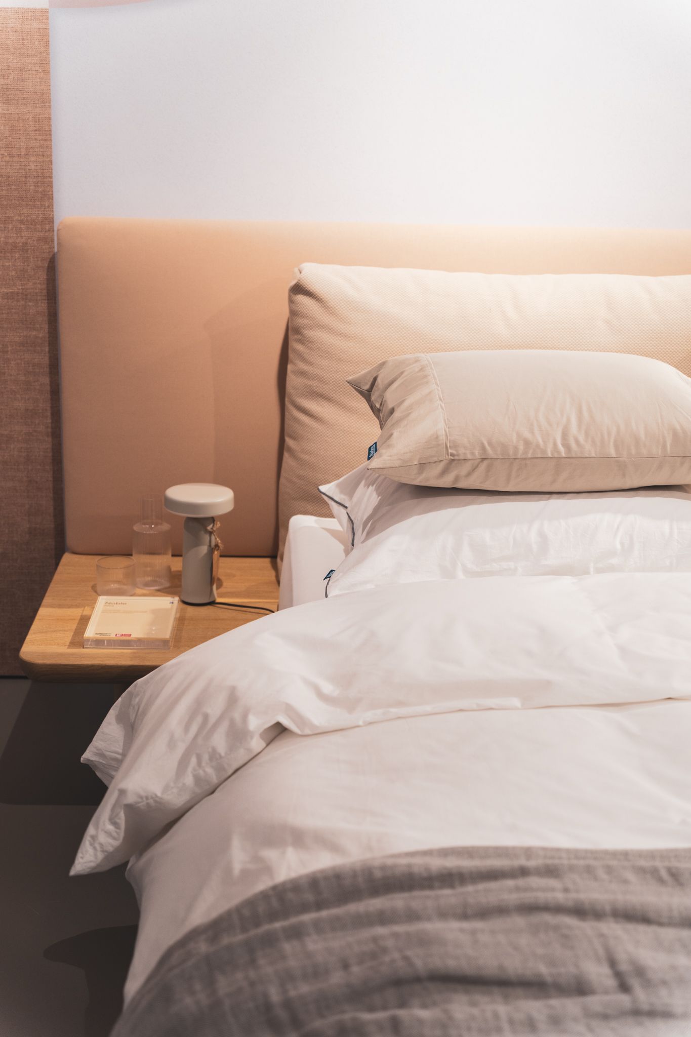 Een modern bed met een gestoffeerd beige hoofdbord, wit beddengoed en een houten nachtplankje.