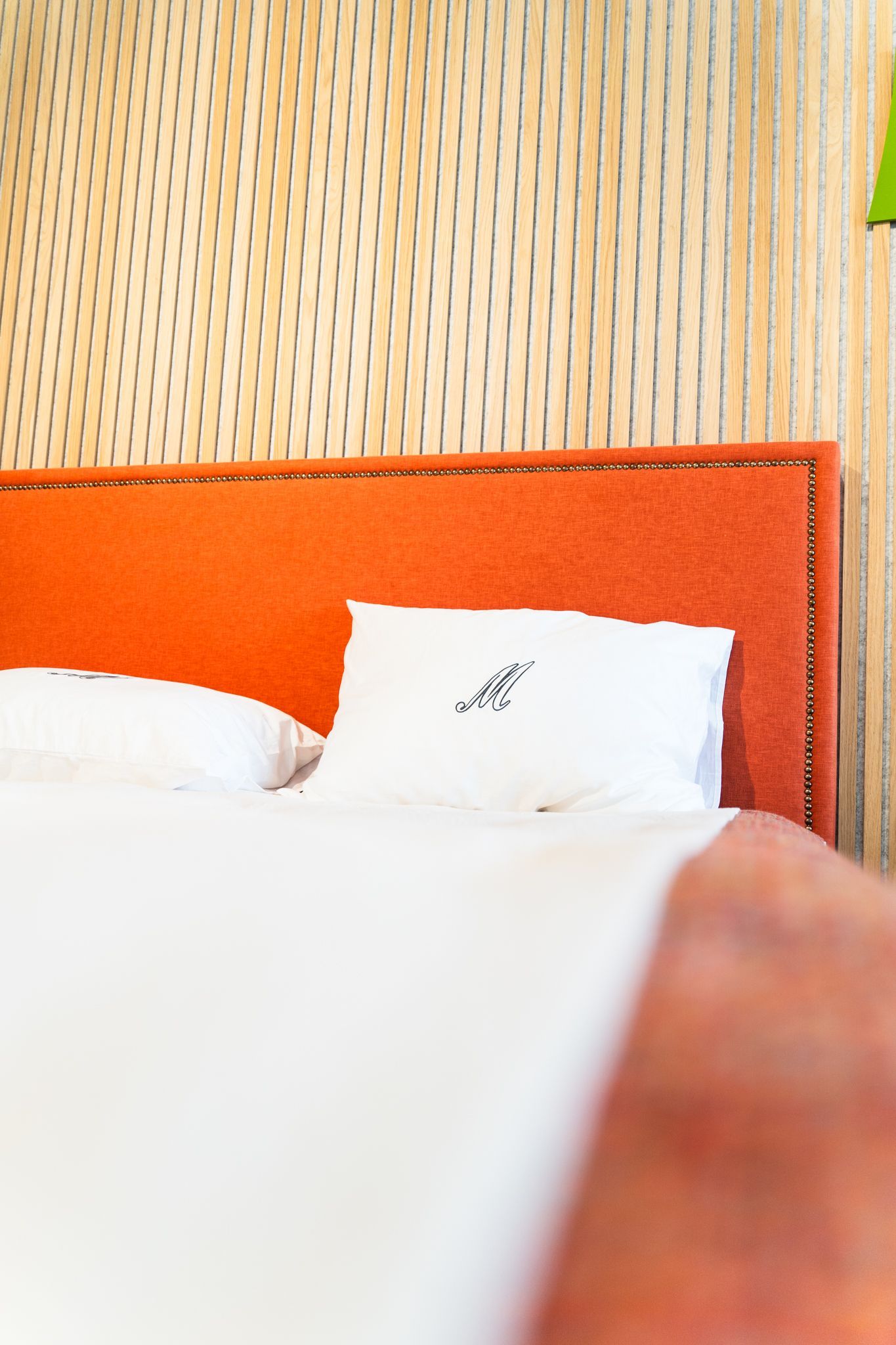 A bed with white pillows, one featuring a small logo, set against an orange headboard and a textured patterned wall.
