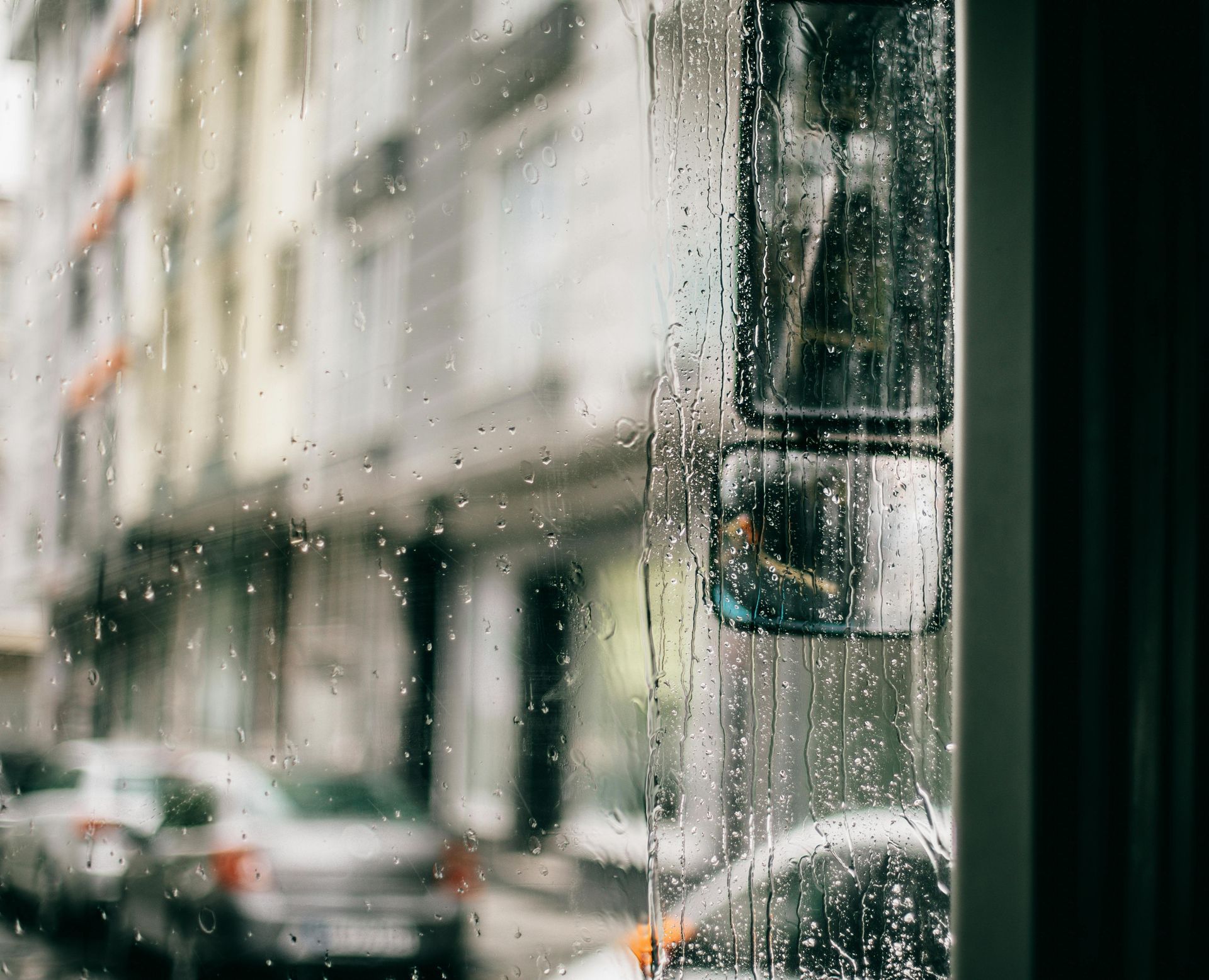 Rainy bus window view of a street, with parked cars and building facades visible through the wet glass.