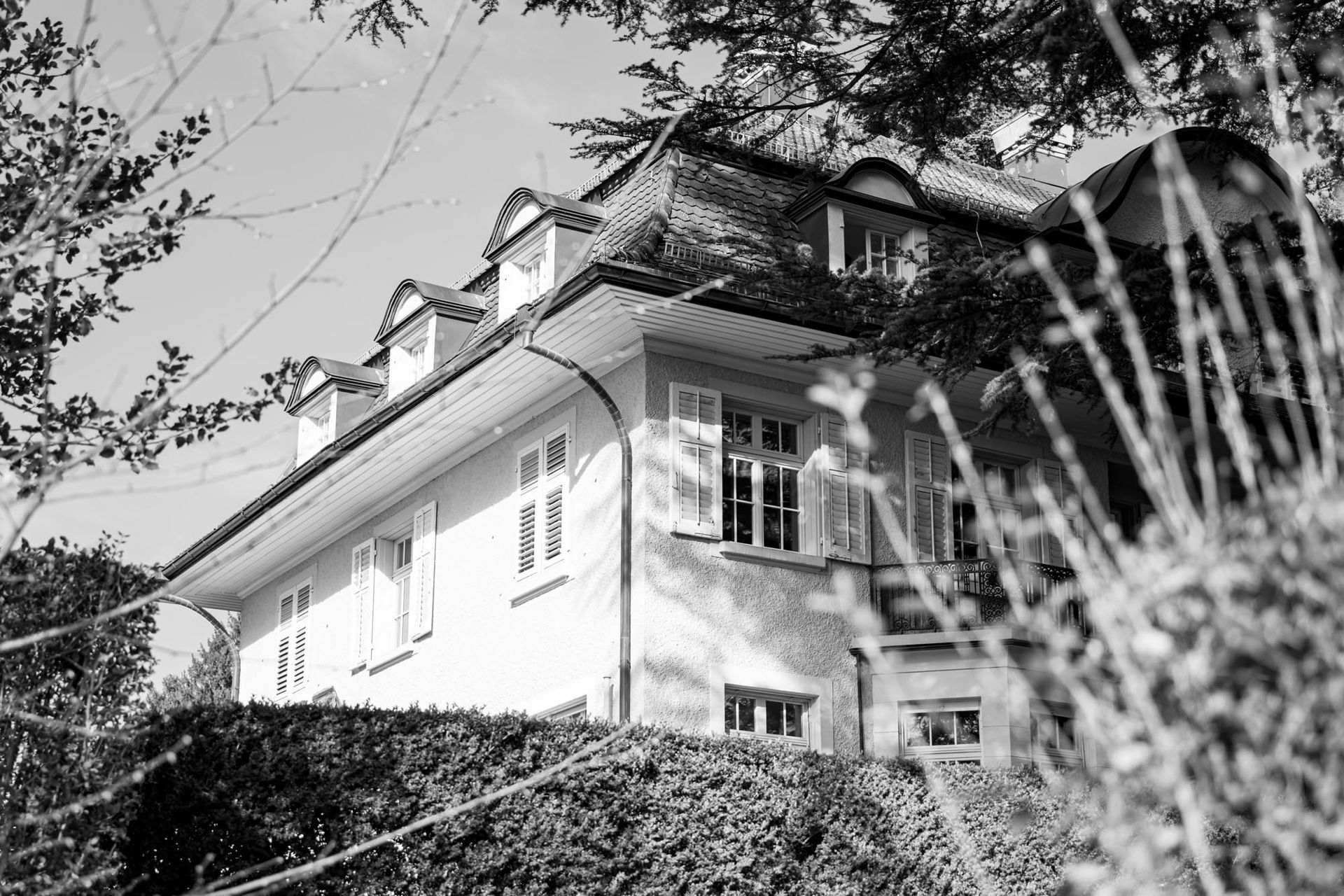 Schwarz-Weiß-Aufnahme eines Hauses am Hang, teilweise eingerahmt von Bäumen und Laubwerk
