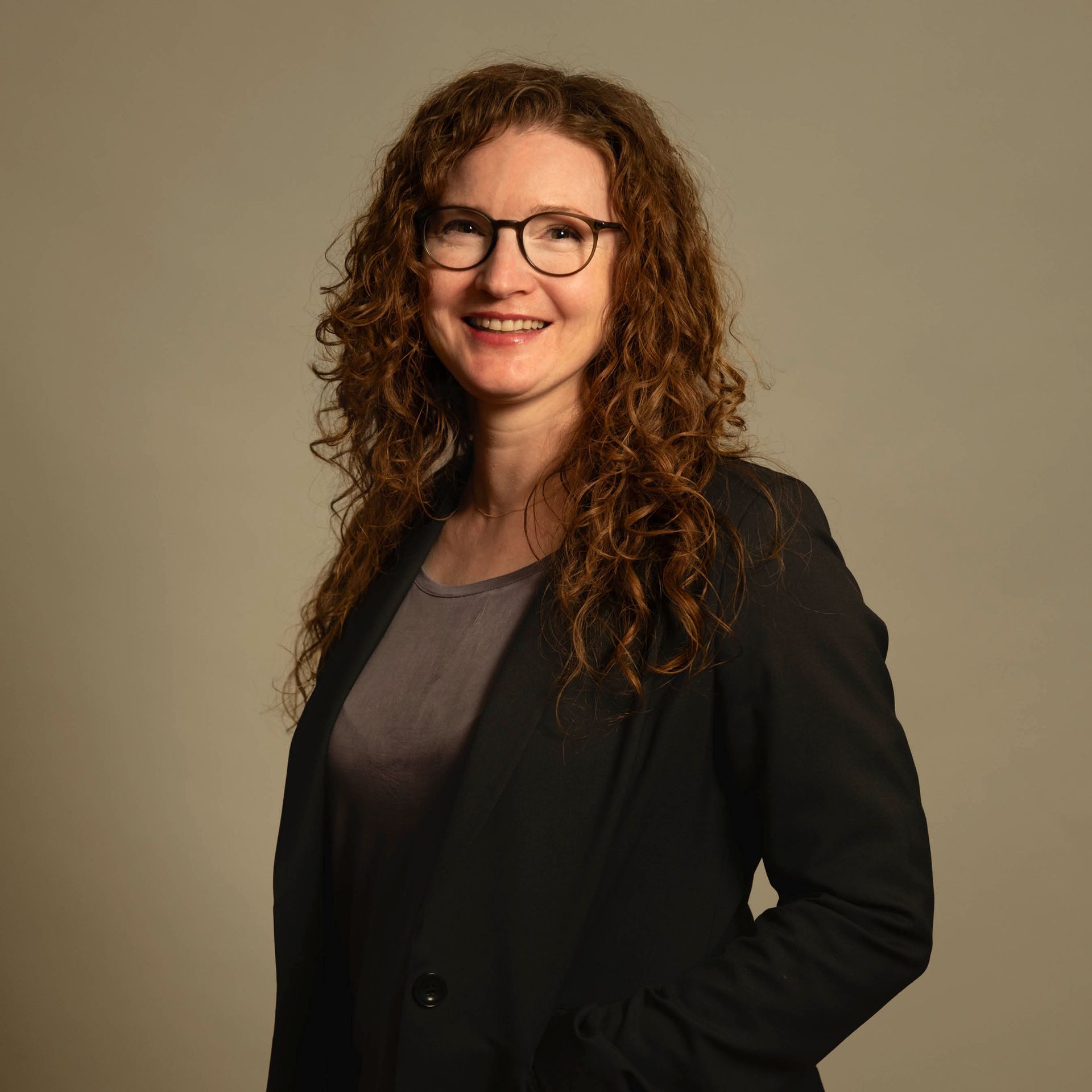 Lächelnde Frau mit lockigem Haar und Brille in einem schwarzen Blazer vor schlichtem beigefarbenem Hintergrund