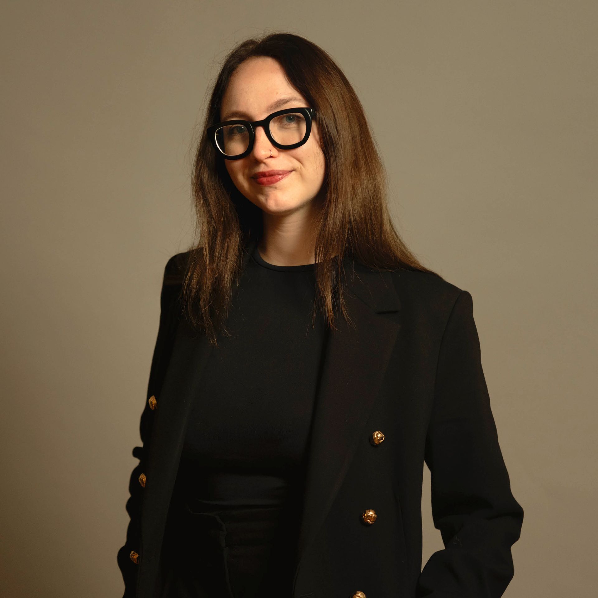 Frau in schwarzem Blazer und Brille, vor einem schlichten beigen Hintergrund.