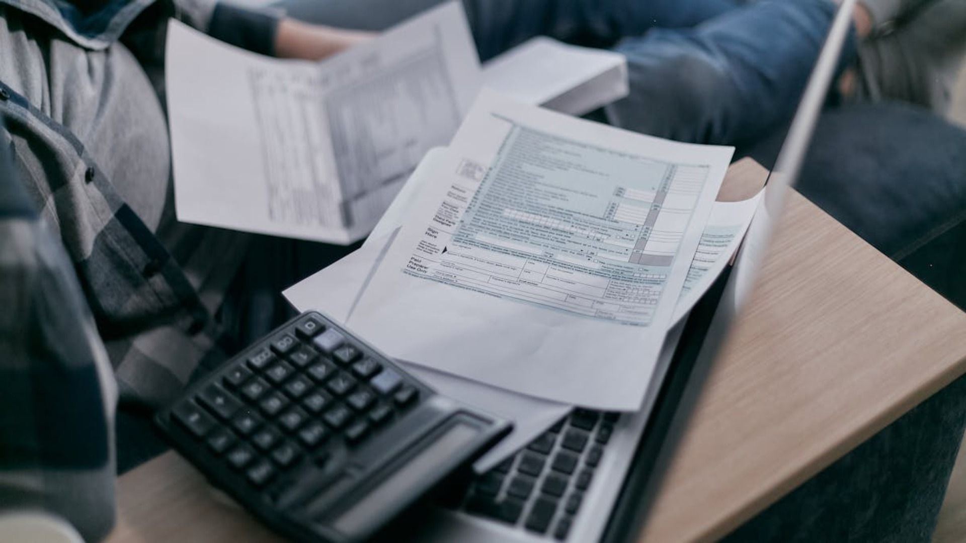 Persona haciendo impuestos, con calculadora, papeles y computadora portátil en su regazo.