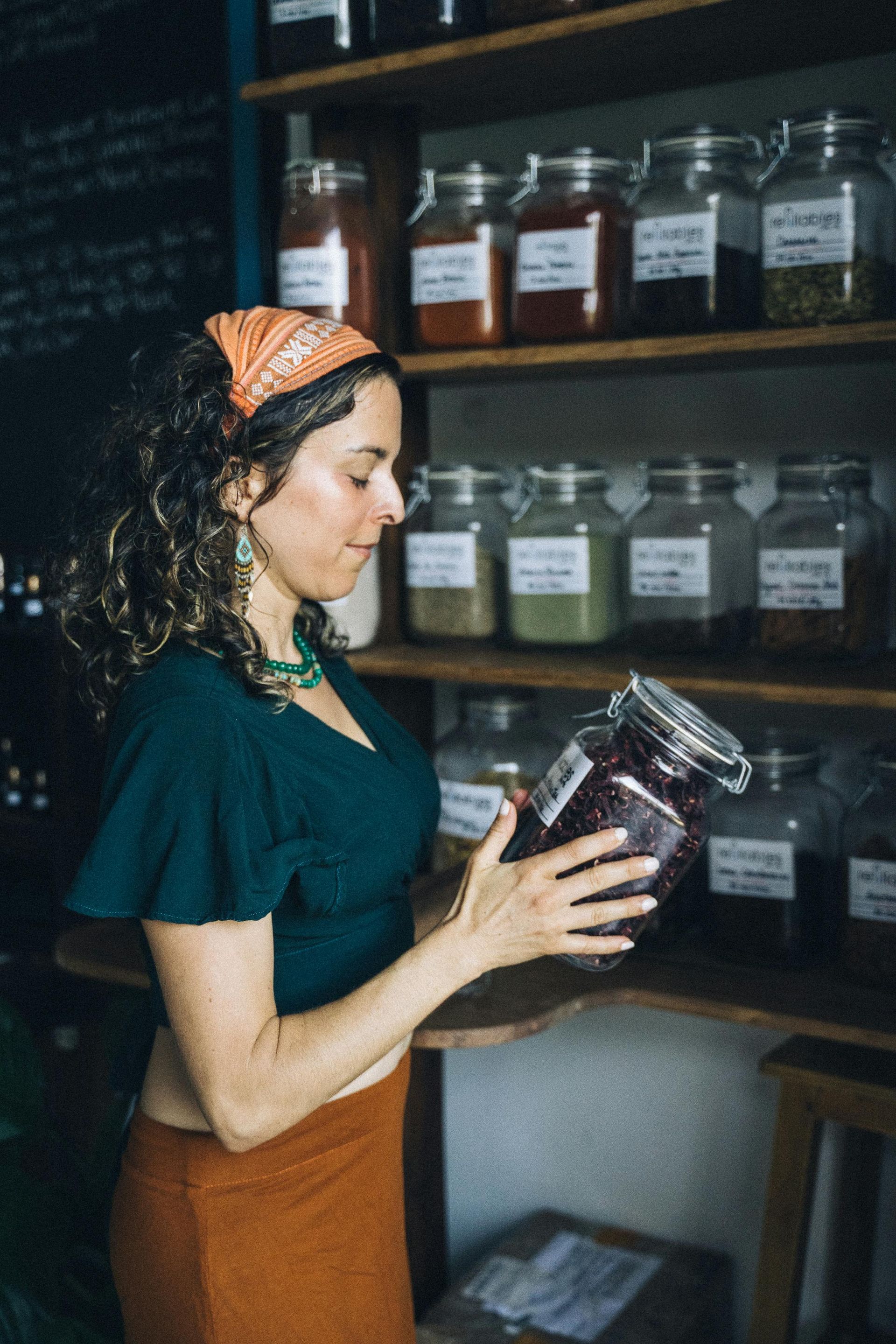 Woman examining a jar of herbs in a shop with shelves of spices. She's wearing a green top, orange skirt and headscarf.