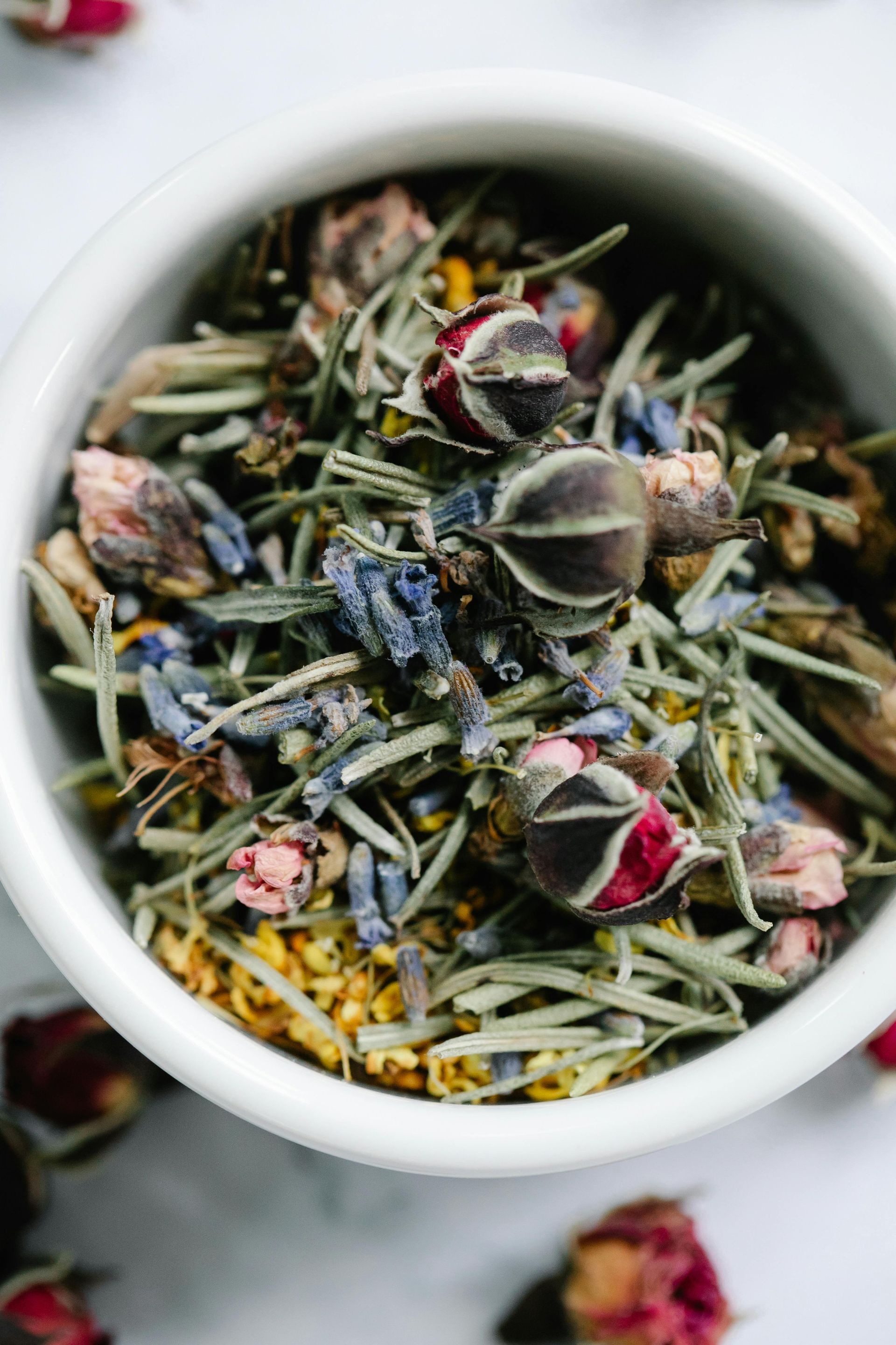 White bowl filled with dried herbal tea, including rosebuds, lavender, and green leaves.