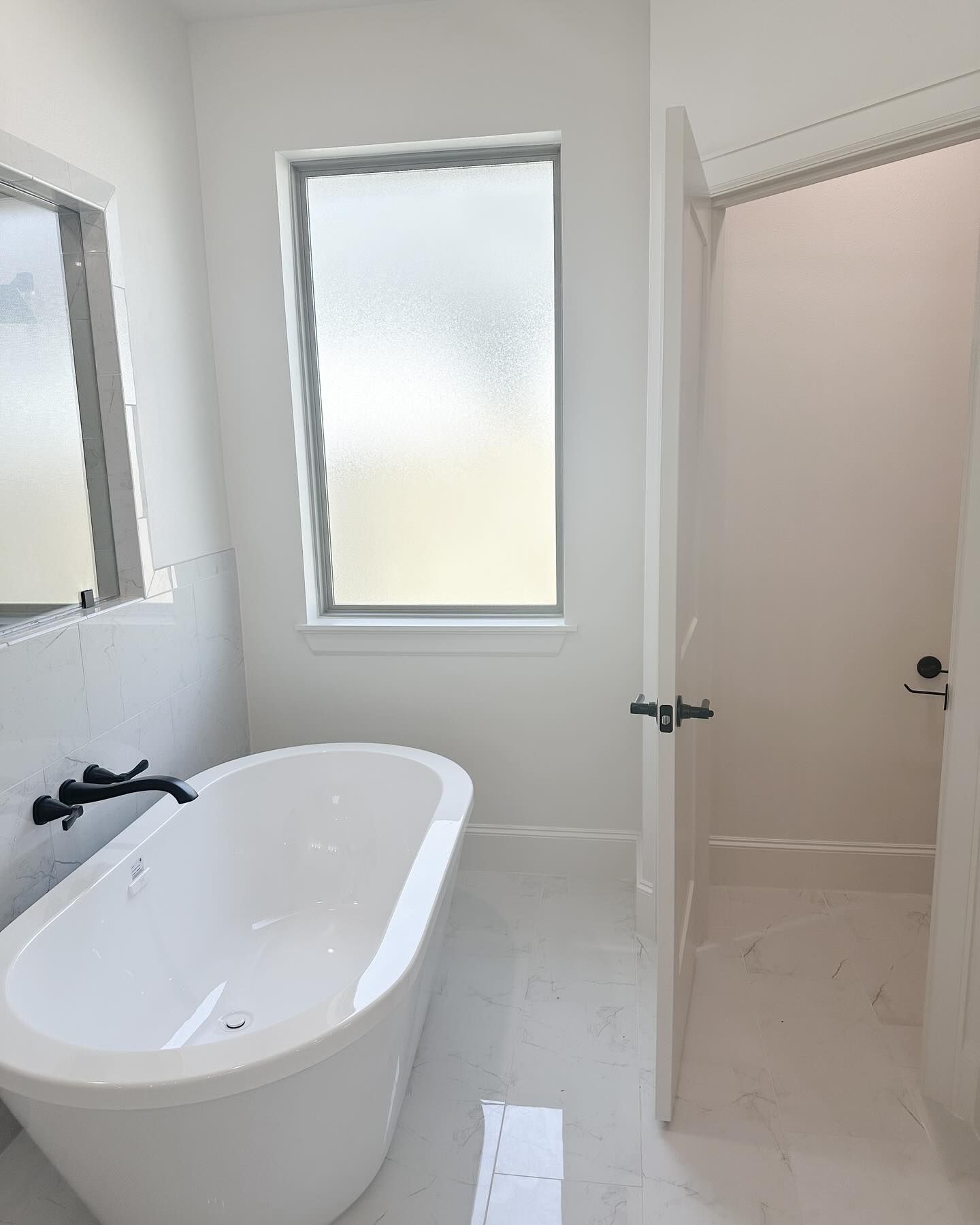White bathroom with a freestanding tub, frosted window, and an open door.