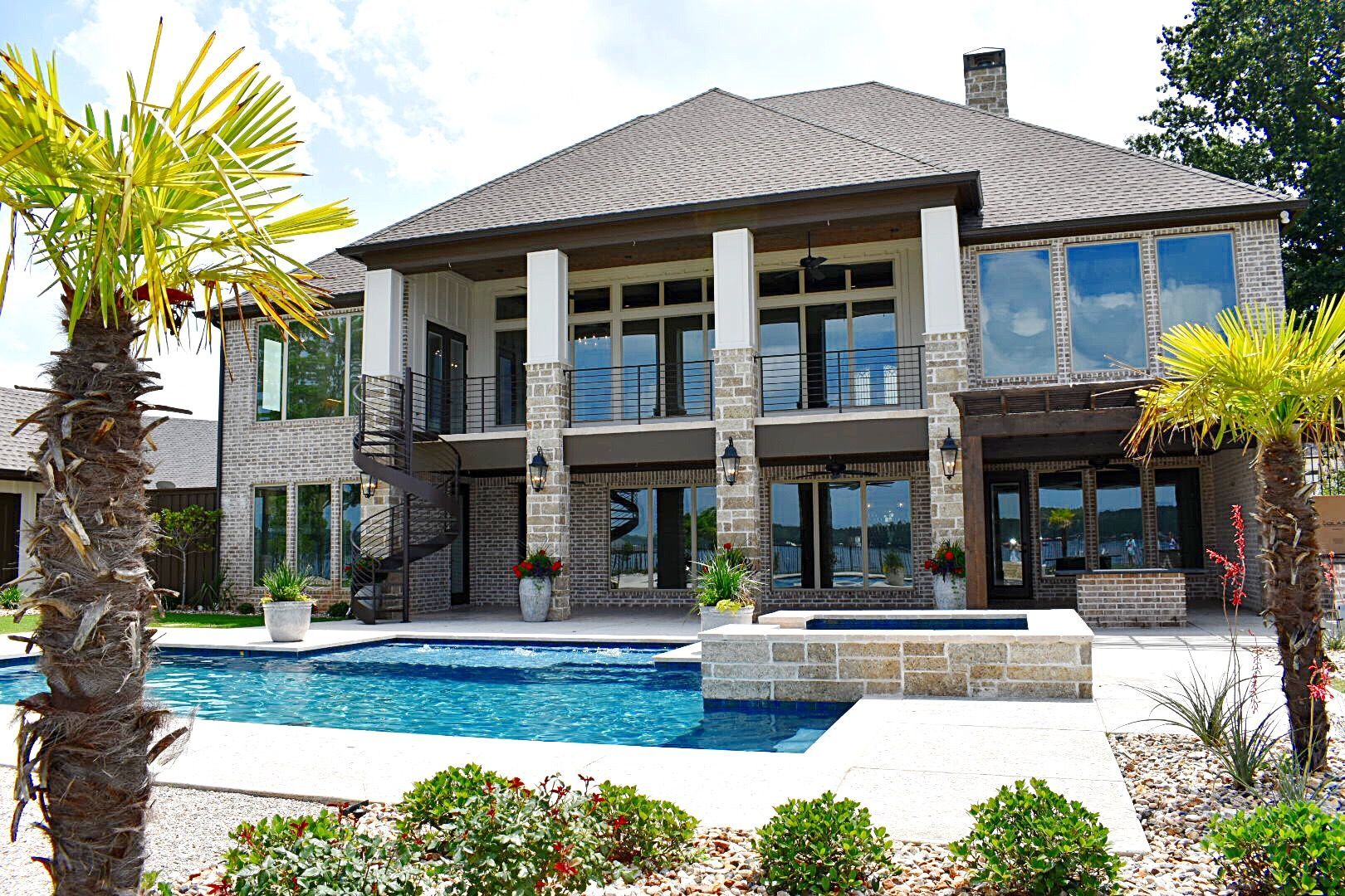 Luxurious two-story house with a pool and palm trees, overlooking a body of water on a sunny day.