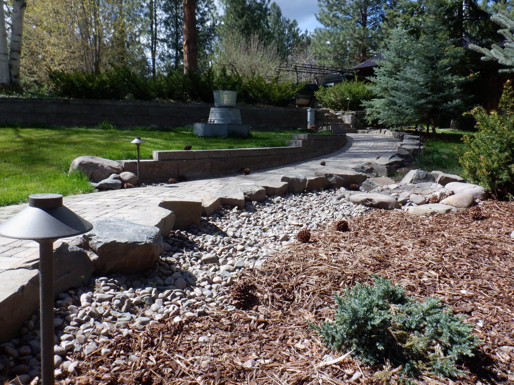 A winding stone path leads through a landscaped yard with rocks, mulch, and small trees.