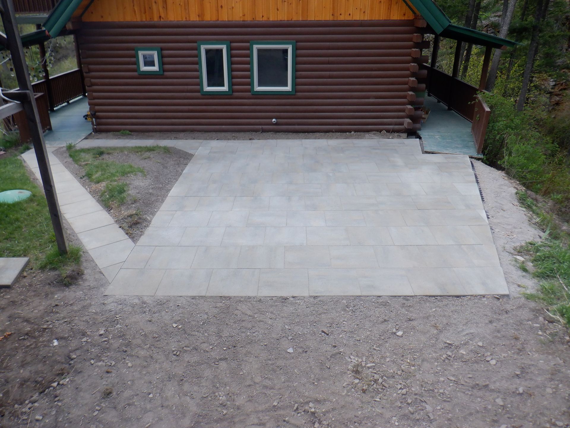 A concrete patio and walkway in front of a log cabin with two windows and a ramp to the right.