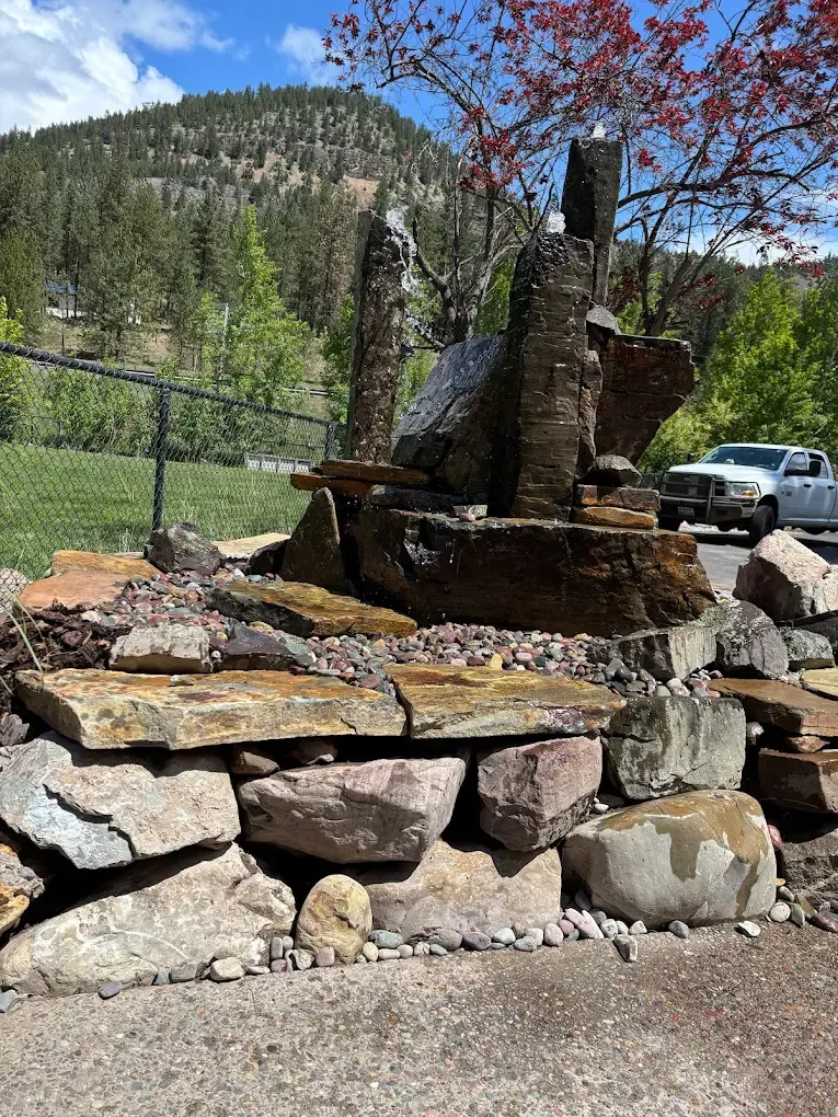 Stone fountain with stacked rock base; green trees, blue sky, and a mountain backdrop.