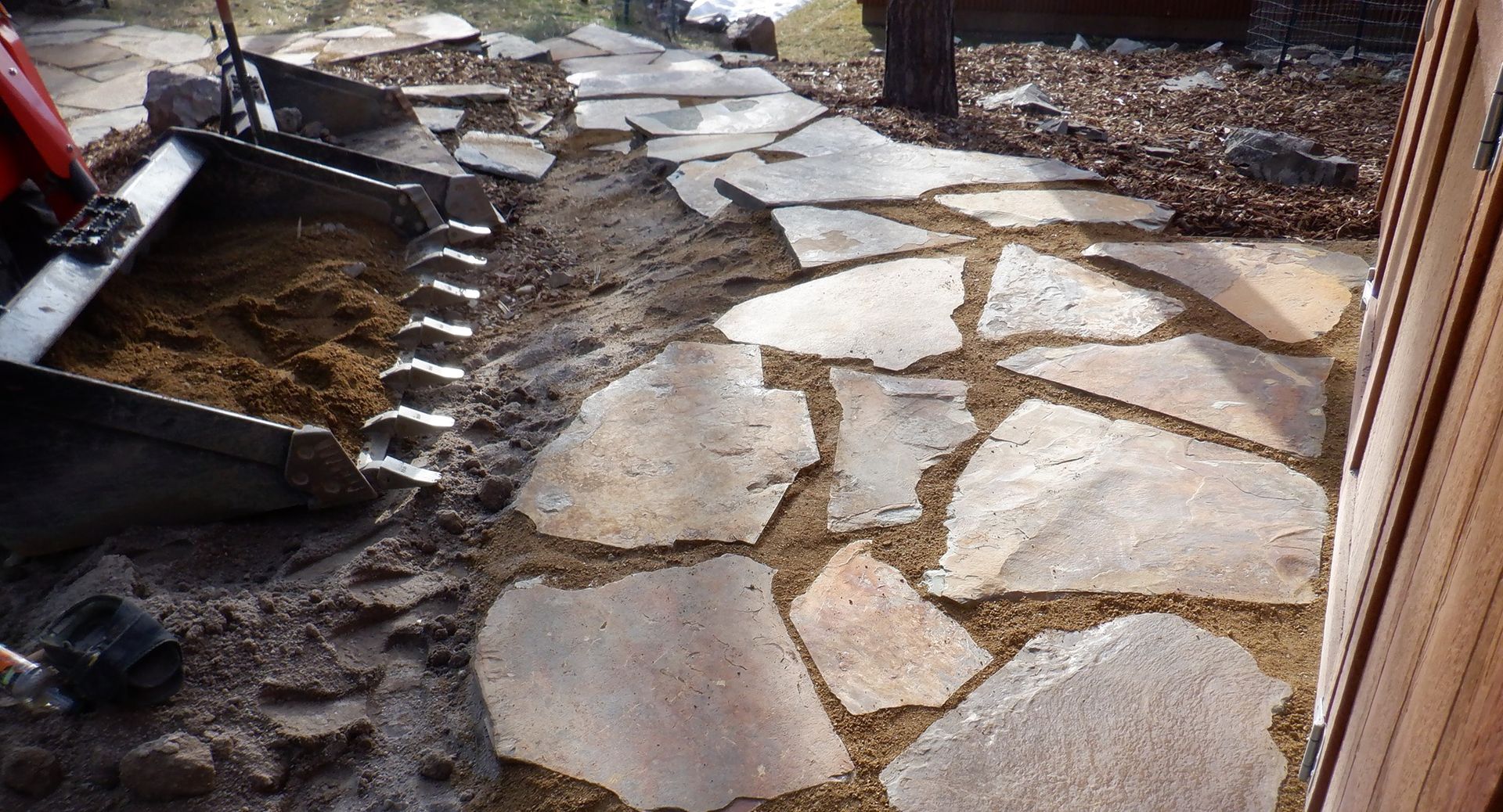 Flagstone pathway under construction; tractor bucket holds material; brown and tan colors.