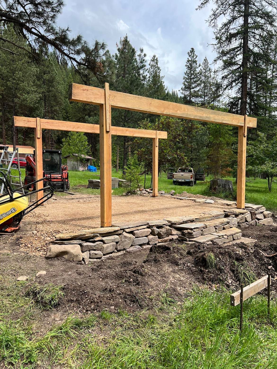 Construction of a wooden pergola with stone base in a grassy outdoor setting.