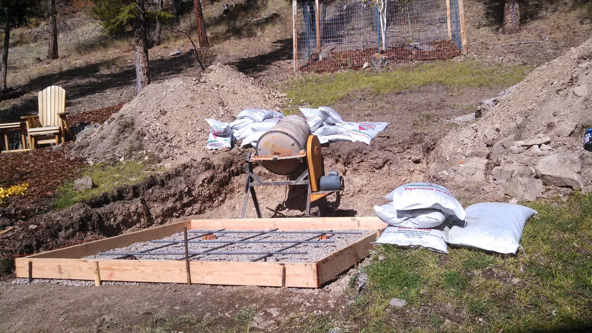 A concrete foundation frame with rebar, cement mixer, and bags of cement on a hillside.