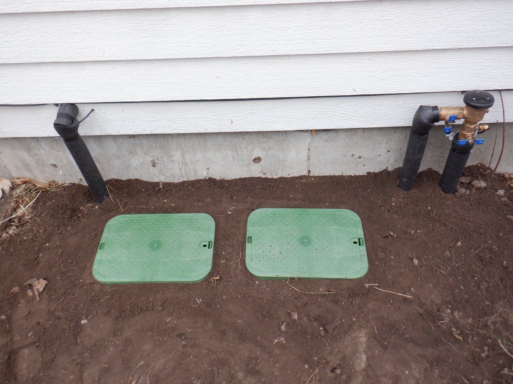 Two green rectangular valve boxes in dirt, near a house with black pipes and a water shutoff valve.