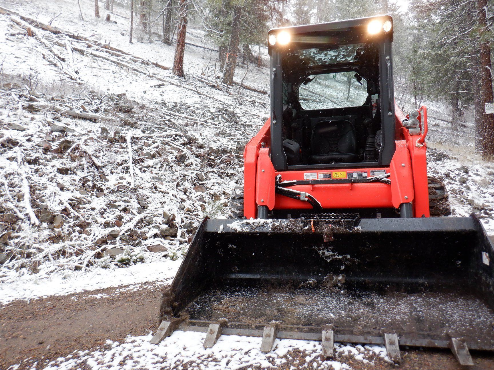 Orange skid steer with bucket on snowy hillside, clearing debris.