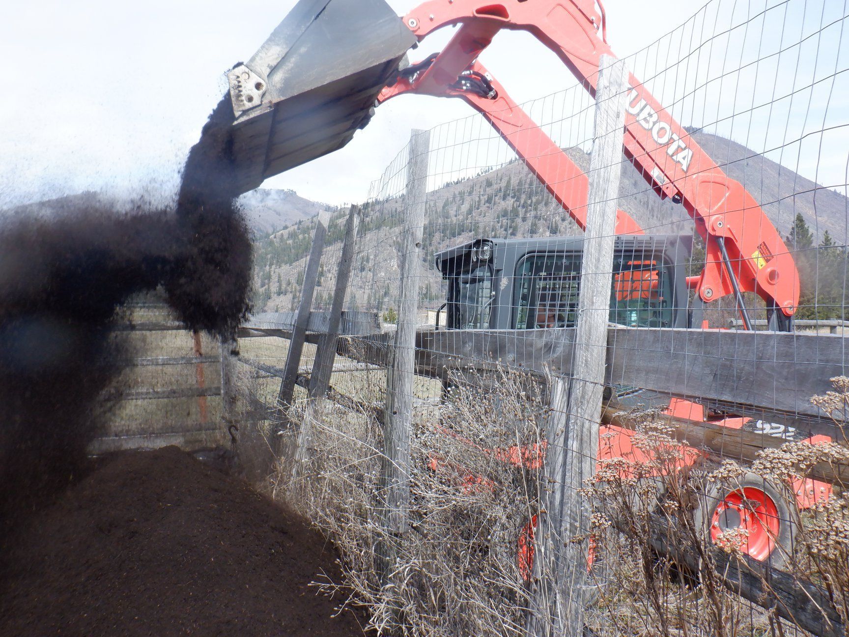 Red excavator dumping dark soil into a fenced area; mountain backdrop.