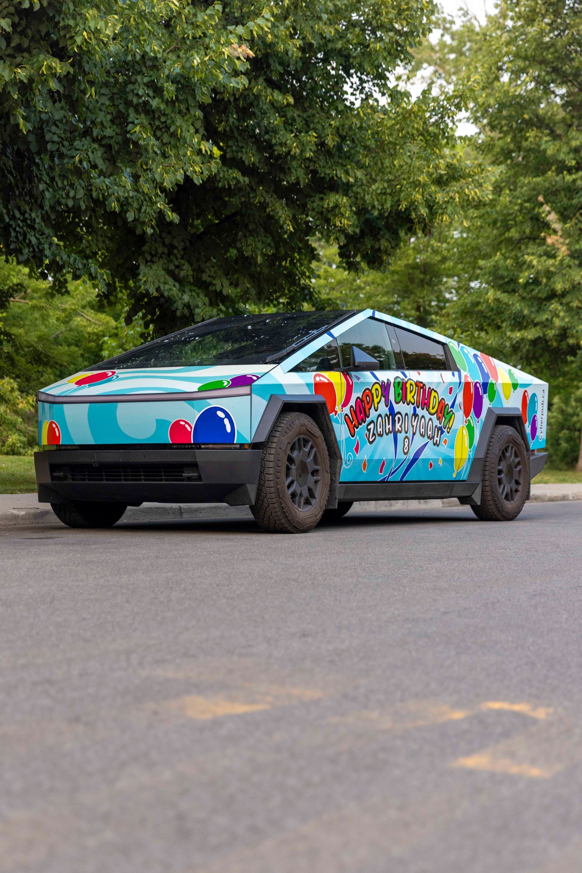 Tesla Cybertruck with colorful artistic wrap parked on a road, trees in the background.