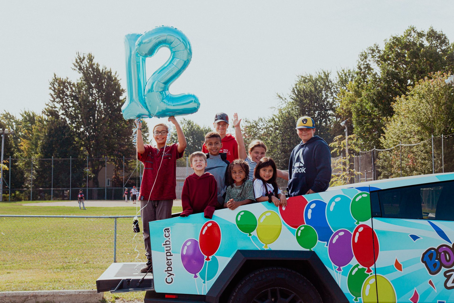 Children on a decorated car holding balloons and a blue 12 balloon; sunny outdoor setting. All standing in a cybertruck birthday.