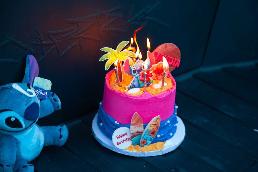 Birthday cake with Stitch theme, lit candles, Stitch plush. Pink, blue, tropical decor. In the frunk of a birthday cybertruck.
