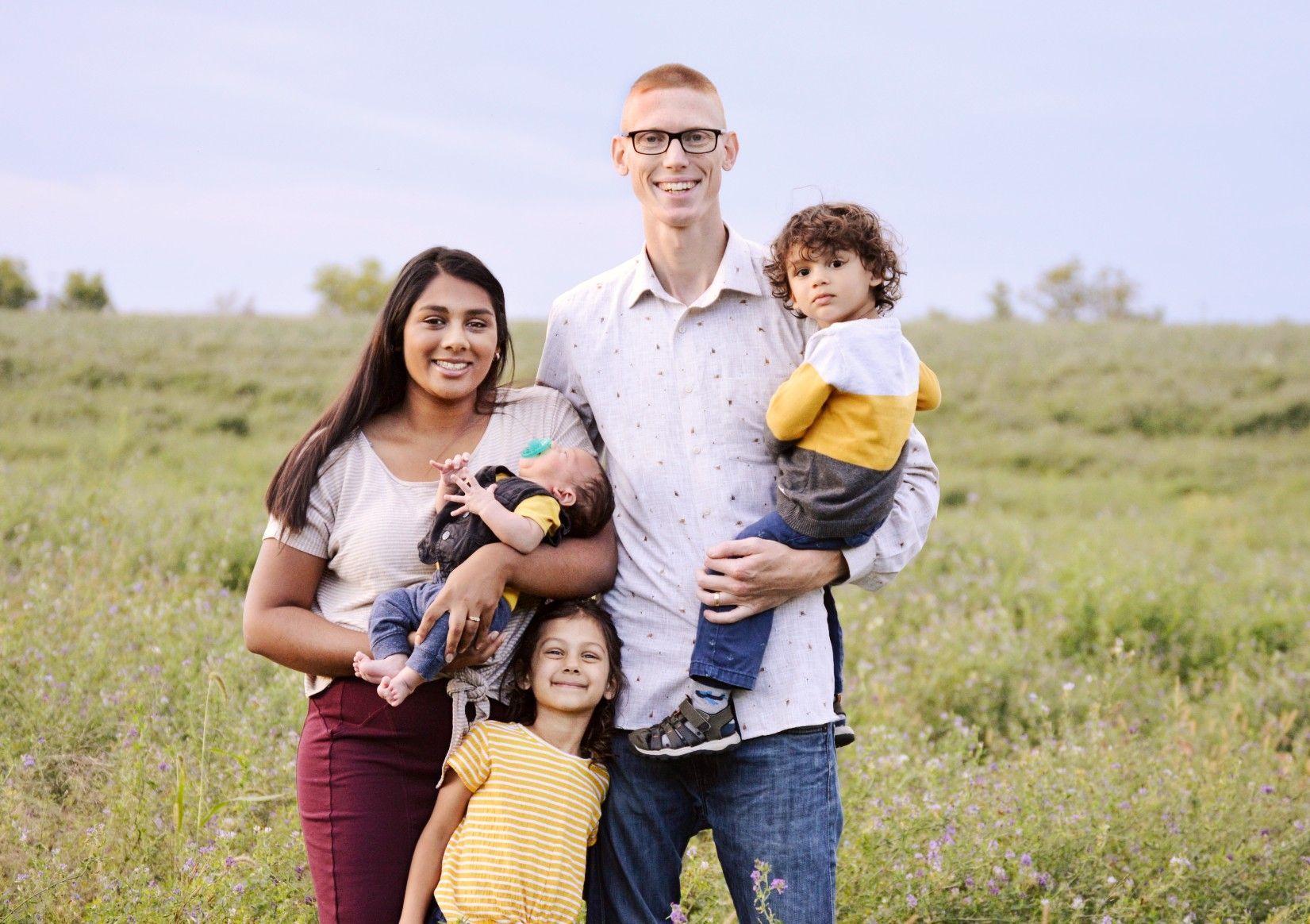 Family of five smiles in a field. Mom holds baby, dad holds toddler, older girl stands in front.
