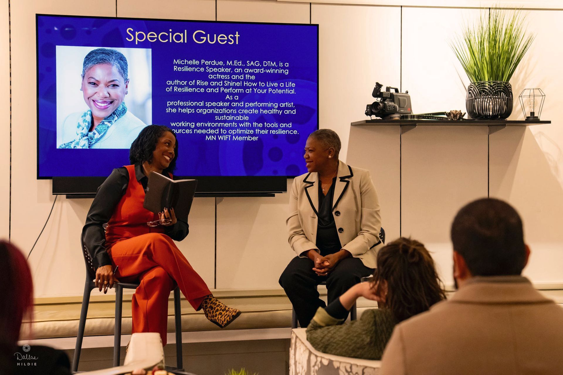 Two women are sitting in front of a television talking to a group of people.